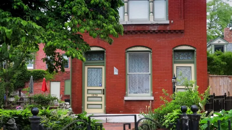 A Red Brick House with Two Green Doors and Lace Curtains, Surrounded by Plants, in St. Louis