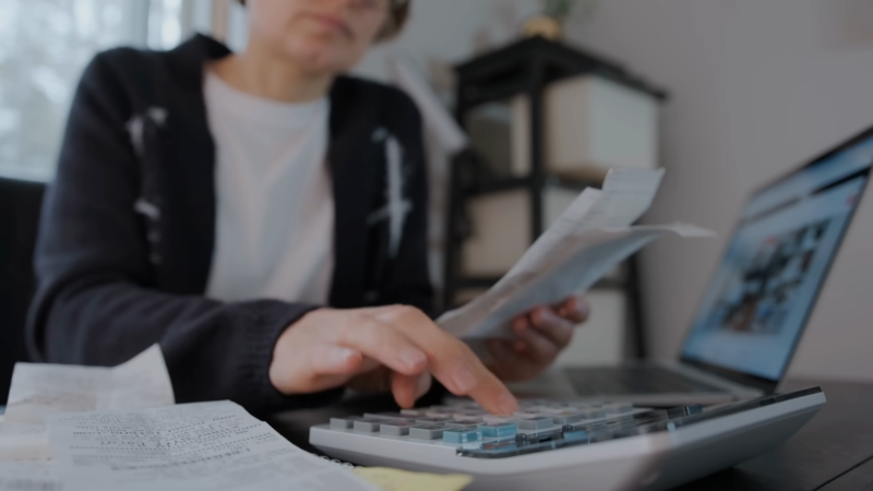 A Woman Examines Financial Documents and Calculates Budget on A Calculator While Using a Laptop, Indicating a Detailed Analysis of Household Income in Alaska