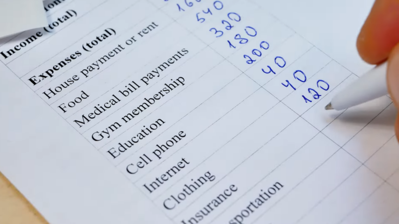 The Image Shows a Close-Up of A Hand Filling out A Budget Sheet Listing Various Expenses Such as House Payment, Food, Medical Bills, Gym Membership, and More, with Handwritten Numbers Next to Each Item, Representing the Cost of Living Breakdown