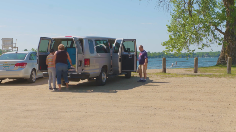 The Image Shows a Group of People Unloading Items from A Van Parked Near a Lakeside on A Sunny Day, Reflecting Community Engagement and Outdoor Activities in Minneapolis