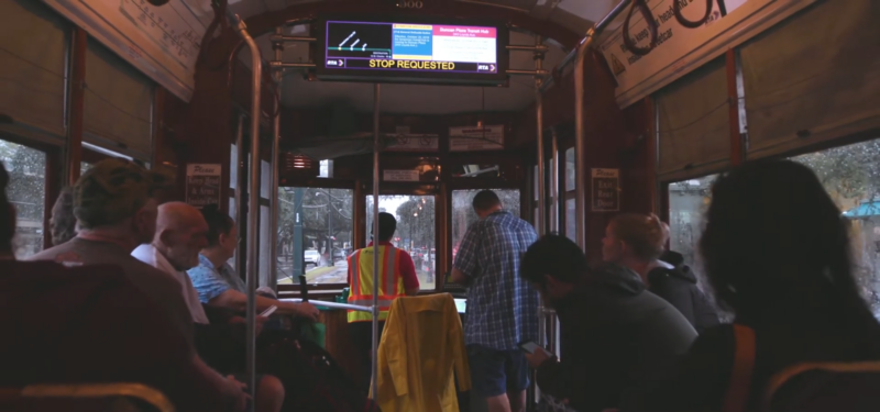 The Image Shows the Interior of A New Orleans Streetcar with Passengers Sitting and Standing, Looking out Of the Windows at The Rainy Street Outside, as One Passenger in A Reflective Vest Prepares to Exit at A Stop