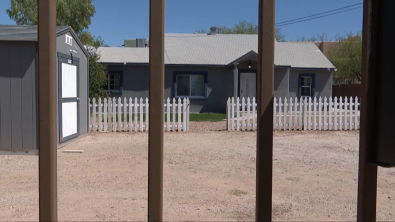 A Gray House with A White Picket Fence, Viewed Through a Brown Wooden Gate, in A Residential Area with A Gravel Yard