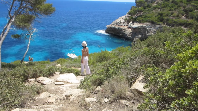 A Woman in A Hat and Summer Attire Stands on A Coastal Path Overlooking the Clear Blue Waters and Rocky Cliffs of Palma De Mallorca, Suggesting a Serene and Scenic Environment for Solo Female Travelers