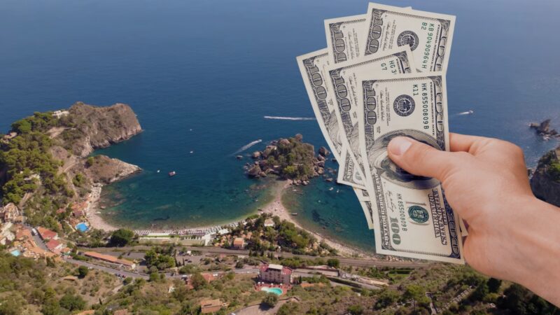 Hand Holding Us Dollars in Front of A Scenic Aerial View of A Coastal Town in Sicily, Illustrating the Article Theme "Is Sicily Expensive?