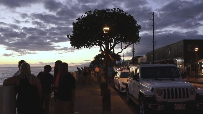 Sunset Street View in Maui County with People Gathered Near the Ocean, Cars Parked Along the Road, and A Tree Lit by A Streetlamp