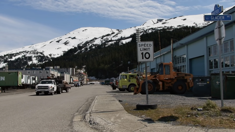 A Street Scene in Whittier, Alaska, with Vehicles and Construction Equipment on Glacier Avenue, Set Against a Backdrop of Snow-Capped Mountains, Highlighting Development Opportunities in The Region