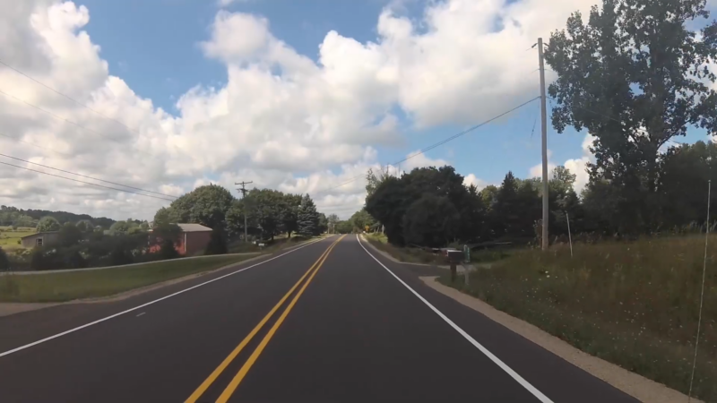 The Image Shows a Rural Road in Michigan, Flanked by Open Fields, Trees, and Scattered Homes Under a Partly Cloudy Sky, Illustrating Typical Countryside Areas Reflecting Trends in Rural Population Distribution