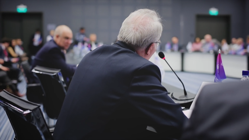 The Image Depicts a Formal Conference or Meeting Setting, Where a Person Is Speaking Into a Microphone, with Other Participants Seated in The Background, Appearing to Be Engaged in Discussion