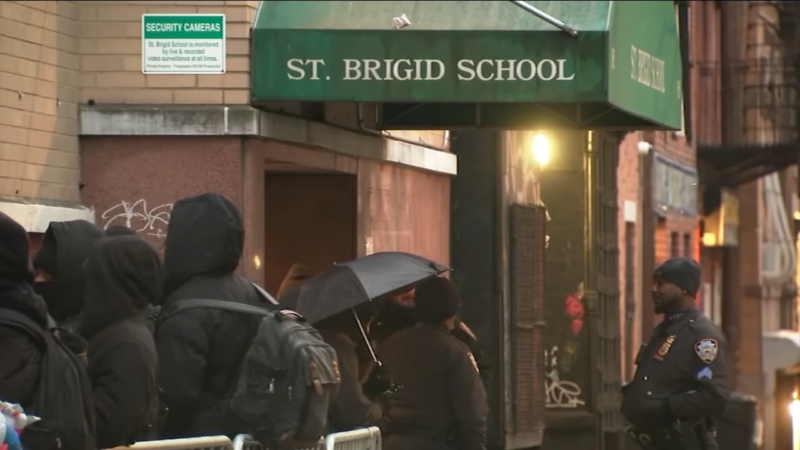 A Group of People, Some Holding Umbrellas and Wearing Hooded Jackets, Stand Outside St. Brigid School in New York City, Indicating the Effects of The Pandemic on Daily Activities and Public Spaces