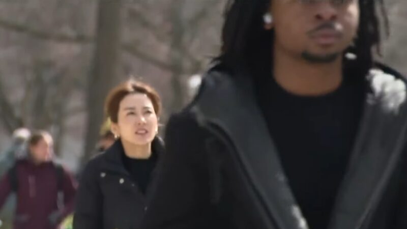 A Woman Walking Outdoors in A Park-Like Setting Is Focused Behind a Man Dressed in Dark Clothing, with Other People Blurred in The Background, Suggesting an Urban Environment in Michigan