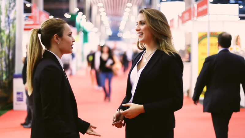 Two Professional Women Engaging in A Discussion at A Busy Business Expo, Exemplifying Workforce Dynamics and Employment Sectors in Boston