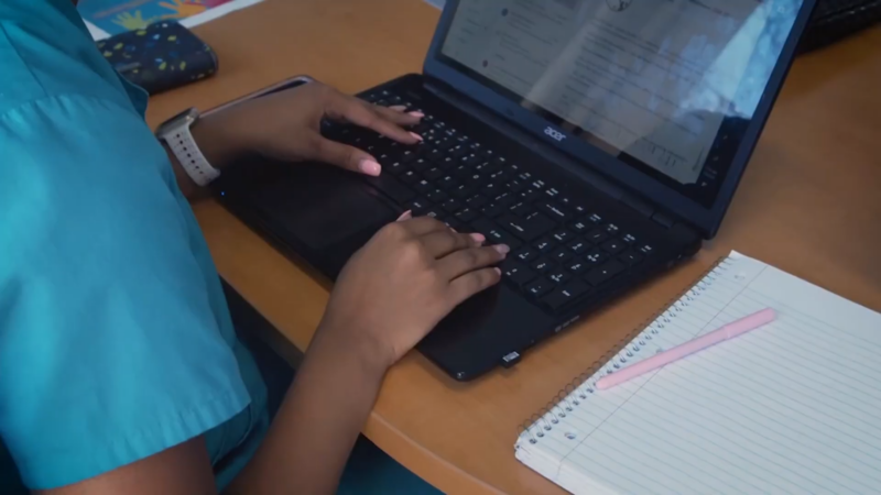 A Nursing Student Types on A Laptop While Taking Notes in An Open Notebook During an Online Education Session