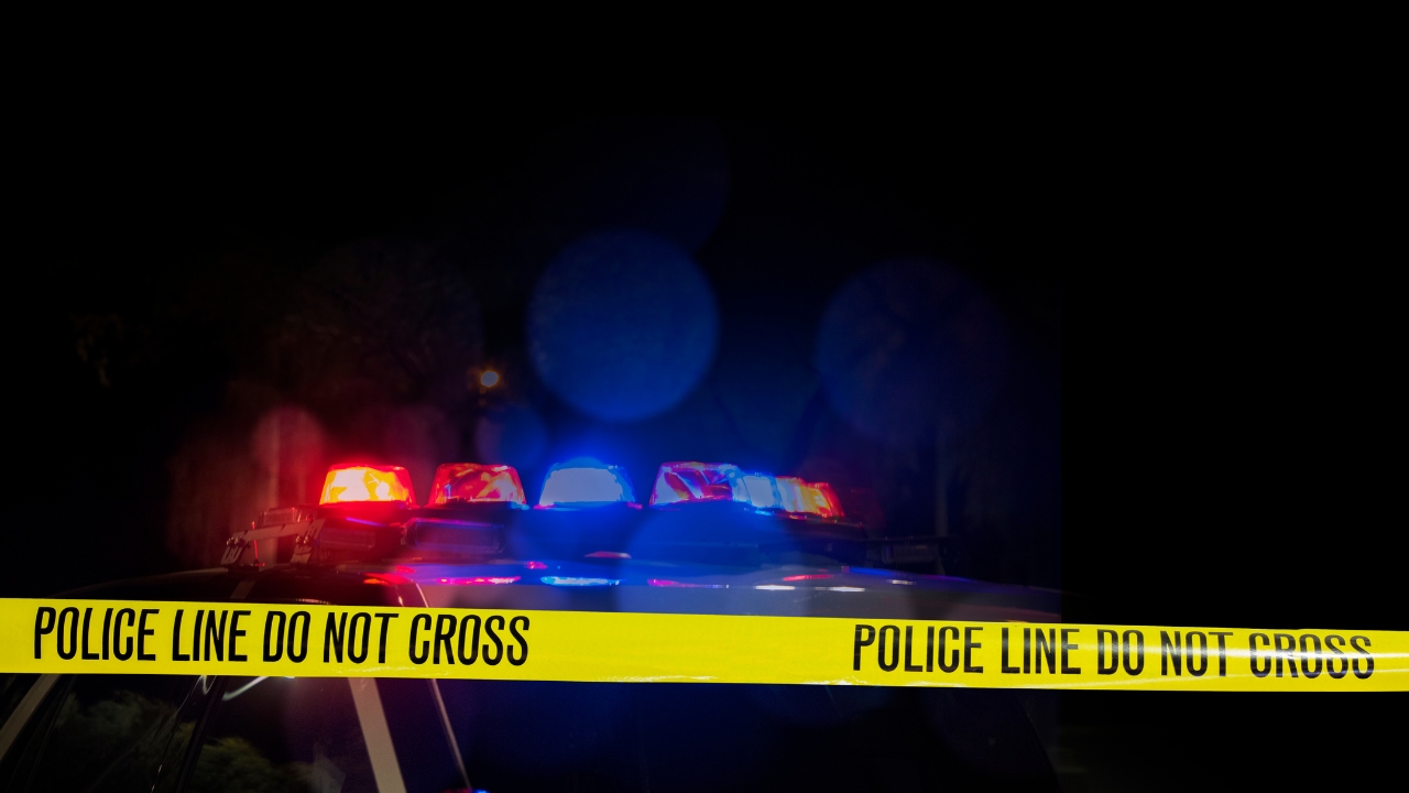 Police tape crosses a dark crime scene with flashing patrol car lights behind it