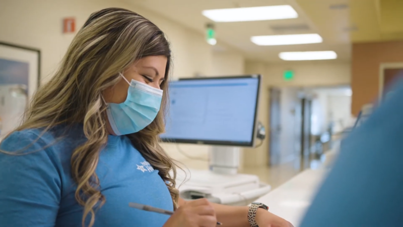 A Healthcare Worker Wearing a Mask and Blue Scrubs Writes Notes at A Workstation in A Medical Facility, Representing Healthcare Services in Texas