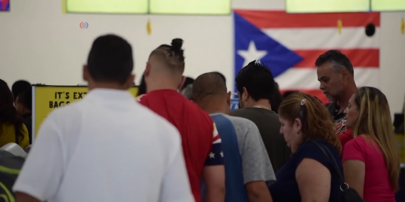 A Group of People Stands in Line at An Airport, with A Puerto Rican Flag Visible in The Background