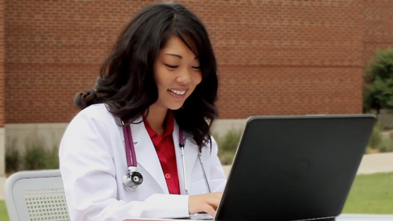 A Smiling Healthcare Professional in A White Coat and Stethoscope Uses a Laptop Outdoors