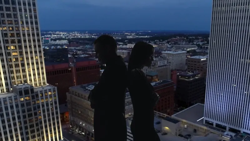 Two People Stand Back-To-Back on A Rooftop at Night, Overlooking the City Lights, with Nebraska's Population in Focus