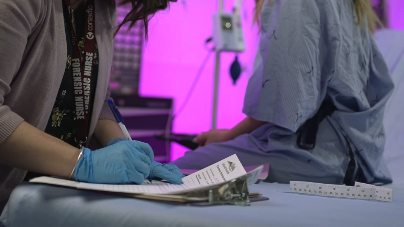 A Forensic Nurse in Gloves Is Documenting Notes While a Patient in A Medical Gown Sits on An Examination Table