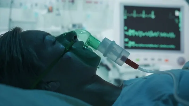 Close-up of a patient lying in a hospital bed with a ventilator mask and an ECG monitor in the background