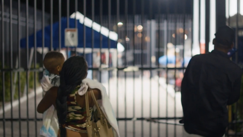 A Woman Carrying a Child Walks Toward a Gated Area at Night