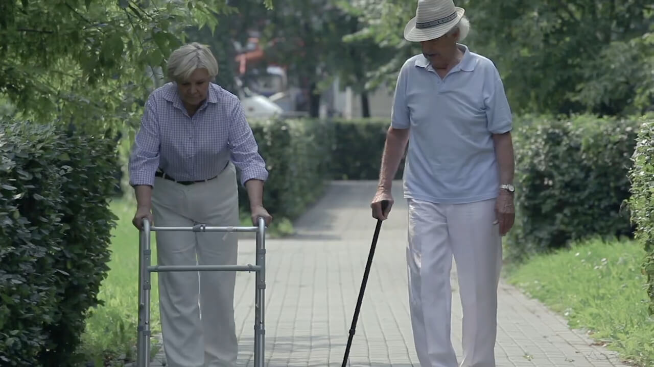 Elderly woman with a walker and elderly man walking on a garden path