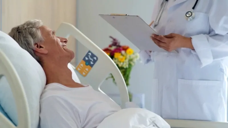 The image shows a patient in a hospital bed speaking with a doctor during a visit
