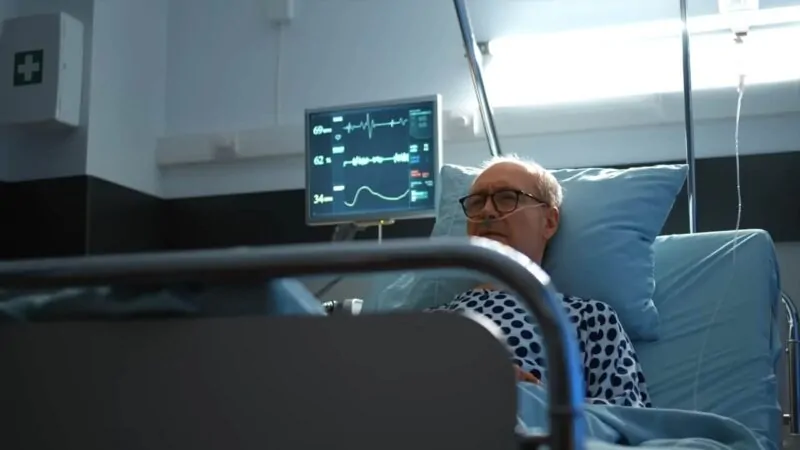 An elderly man wearing glasses and a hospital gown lies in a hospital bed
