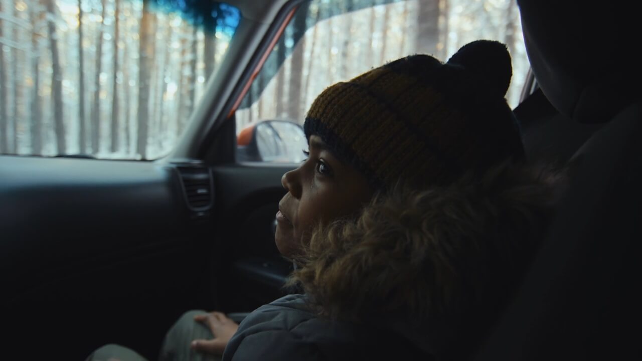 A young child sitting in a car, looking out the window at a forested area with a thoughtful expression