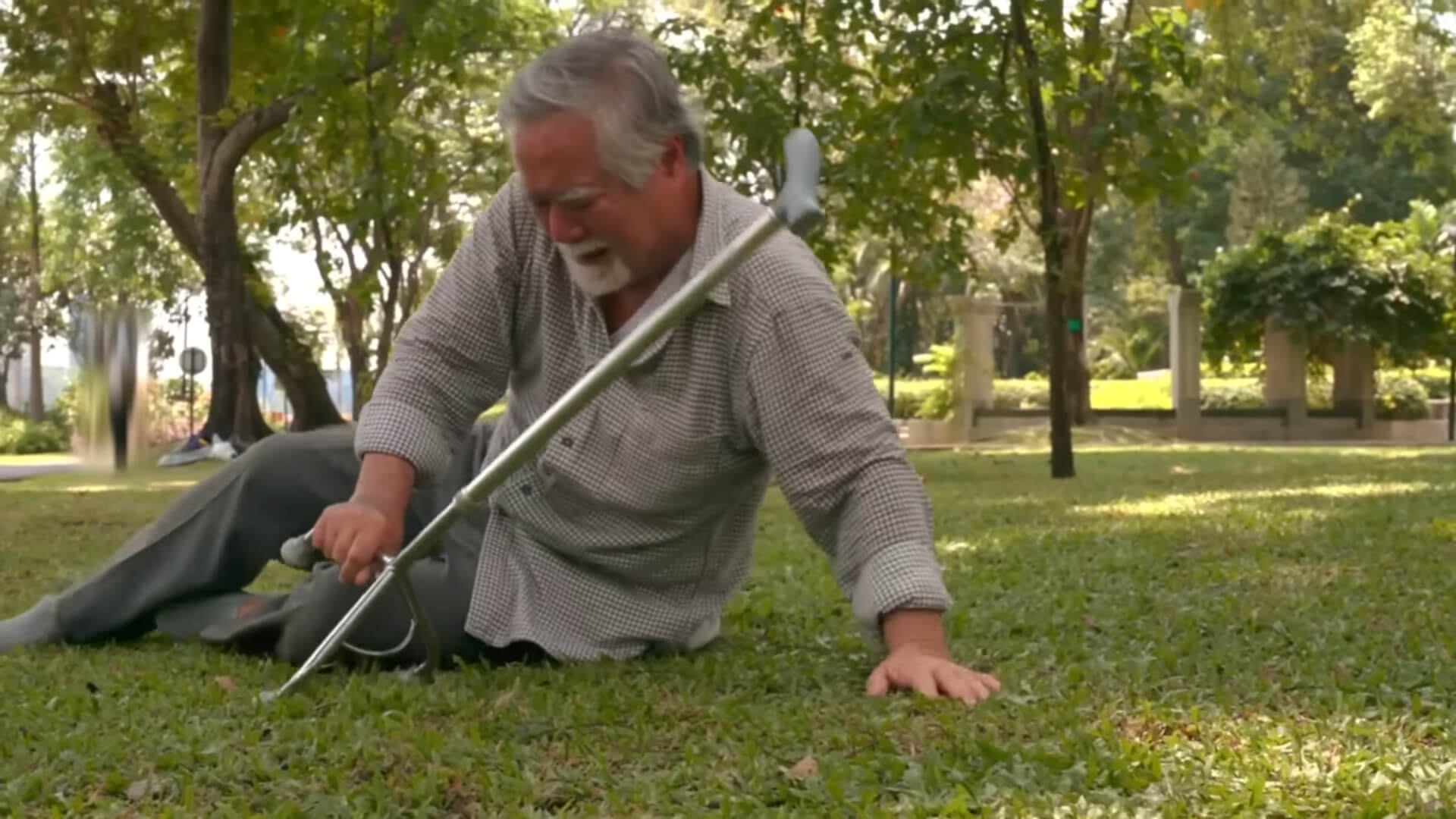 An elderly man sitting on the grass in a park, holding a walking cane and appearing to have fallen