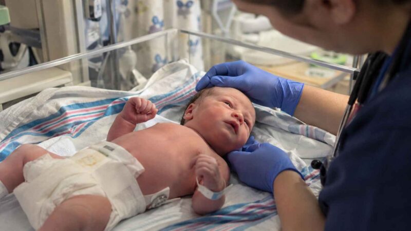 Newborn baby lying on a hospital blanket while a healthcare worker wearing blue gloves examines the head and neck.