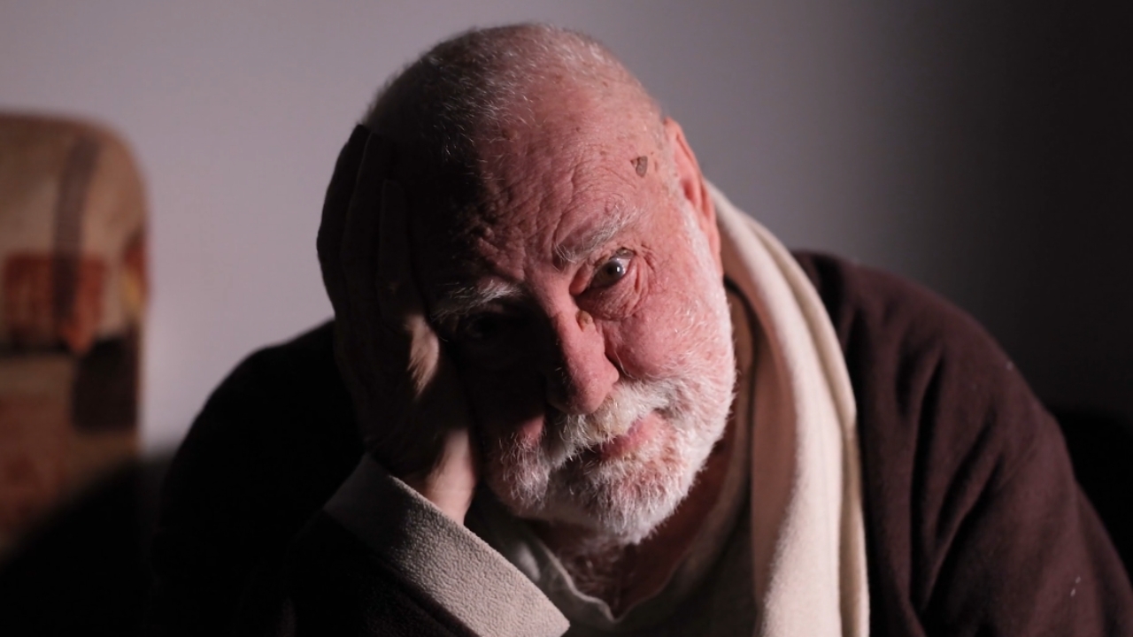 An older man rests his head on his hand in a dim room