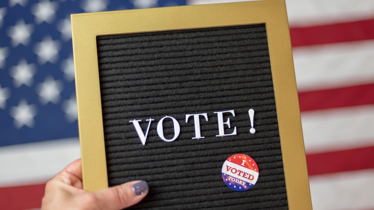 A vote sign and “I Voted” sticker are held in front of an American flag