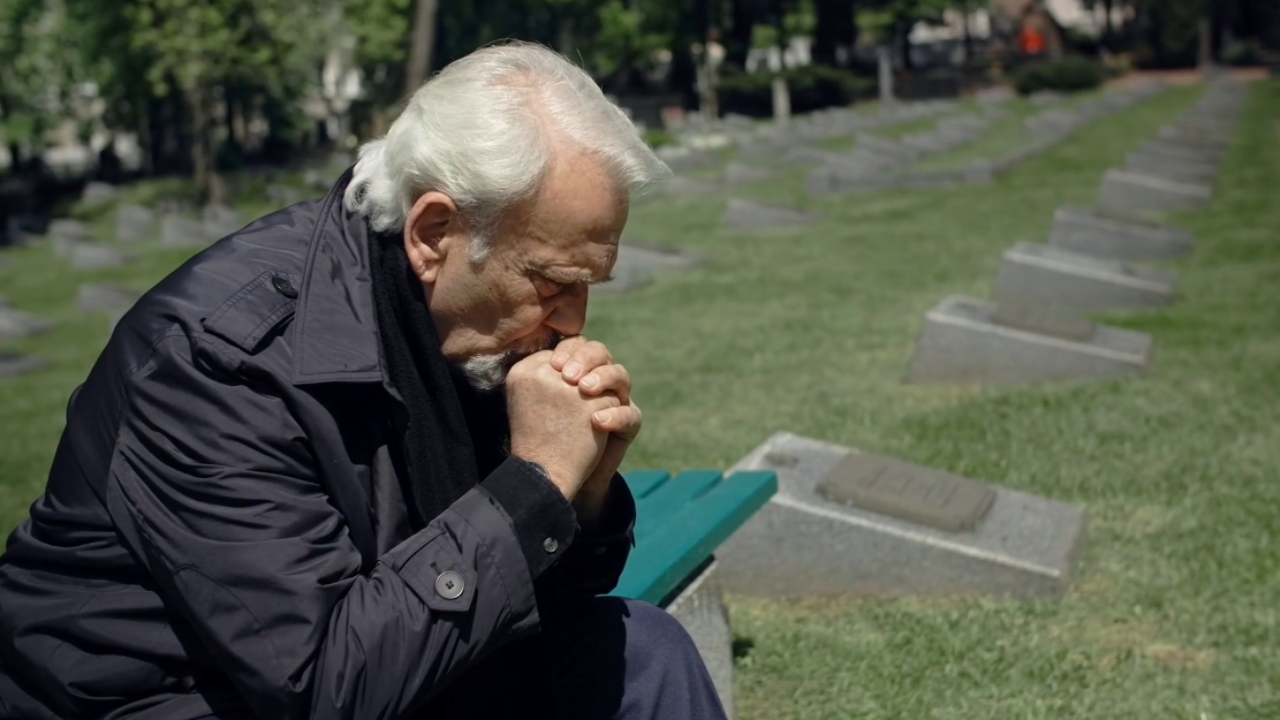 An older man sits beside graves with his hands clasped