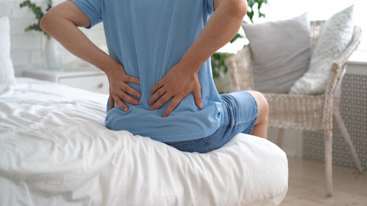 A person sits on a bed and holds their lower back in pain
