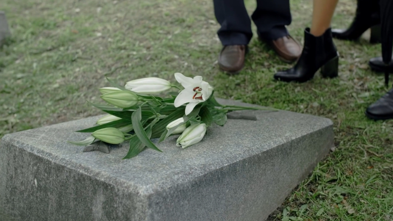 White lilies rest on a gravestone as mourners stand nearby