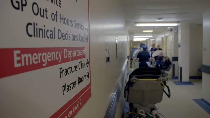 A hospital hallway with a directional sign pointing to the Emergency Department, Fracture Clinic, and Clinical Decisions Unit, with medical staff and patients visible in the background