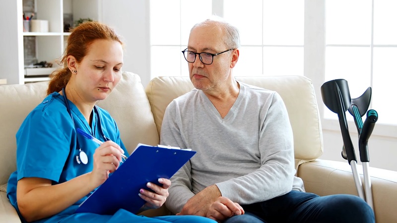 A nurse reviews medical information with an elderly man at home, with crutches nearby, suggesting follow-up care post-ER visit