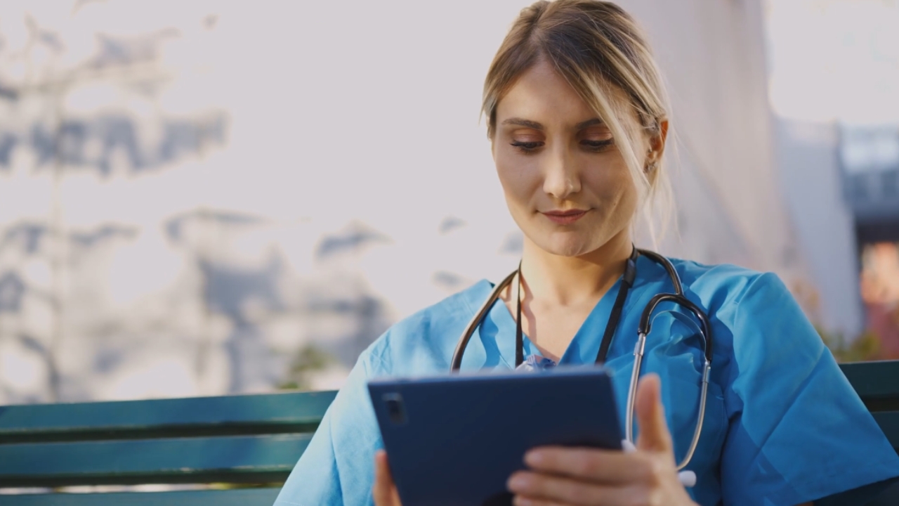 A healthcare worker in scrubs uses a tablet outside