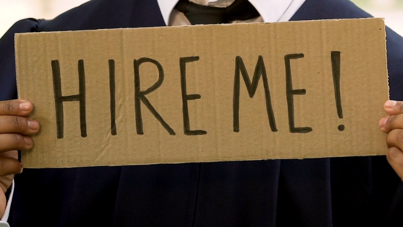 Person holding a cardboard sign that reads "HIRE ME!"