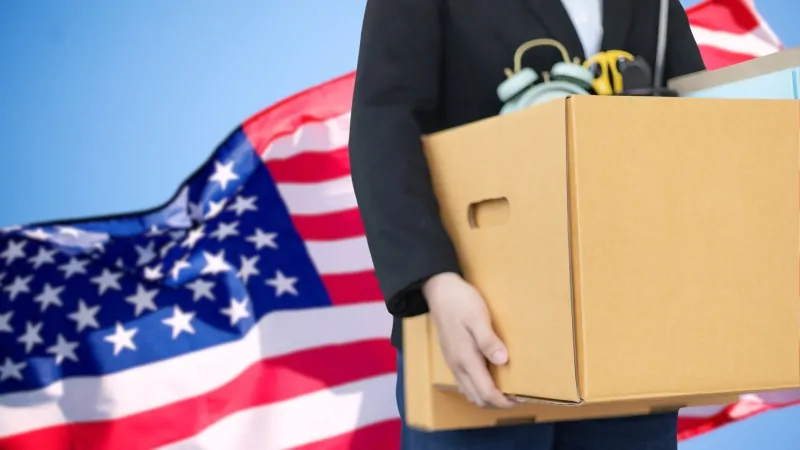 Person holding a box of personal items in front of the American flag, representing job loss and the US unemployment rate