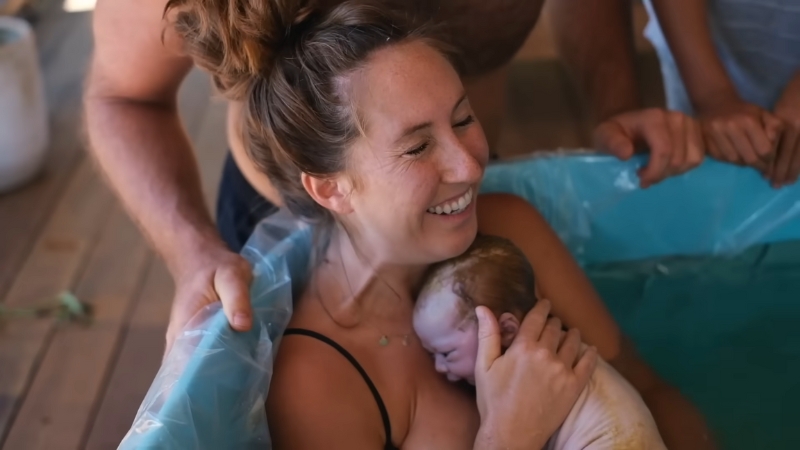 A mother holds her newborn in a birthing tub at home, surrounded by her support team