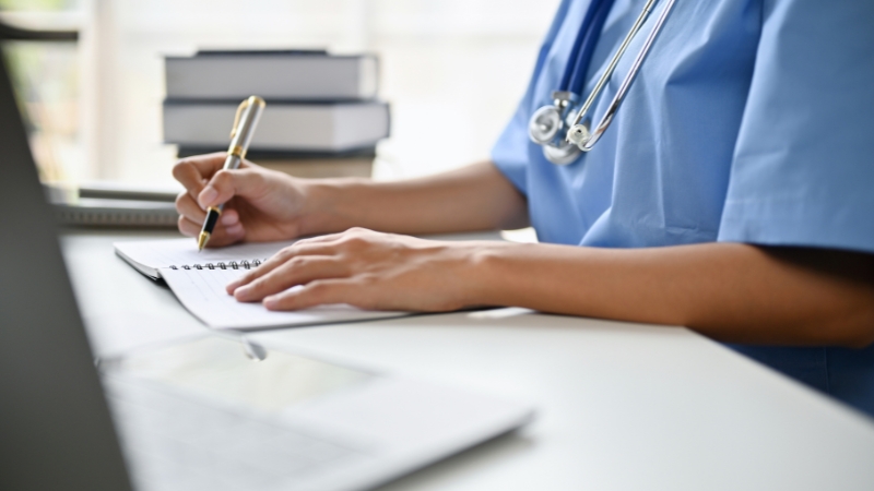 A healthcare professional writes notes at a desk with a laptop and books while planning a private medical practice