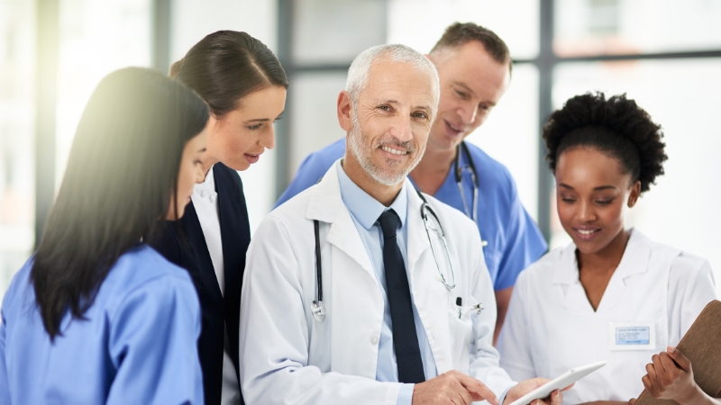 A team of doctors and nurses gathers around a tablet to discuss staffing and equipment needs for a private medical practice
