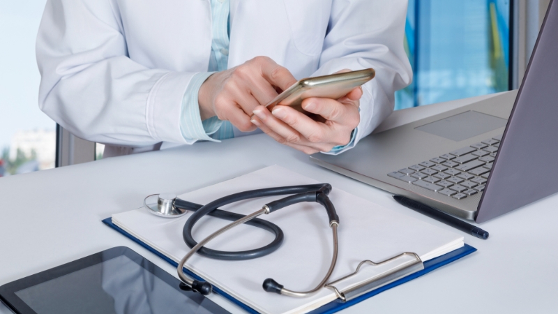A doctor uses a smartphone at a desk with a stethoscope, clipboard, and laptop