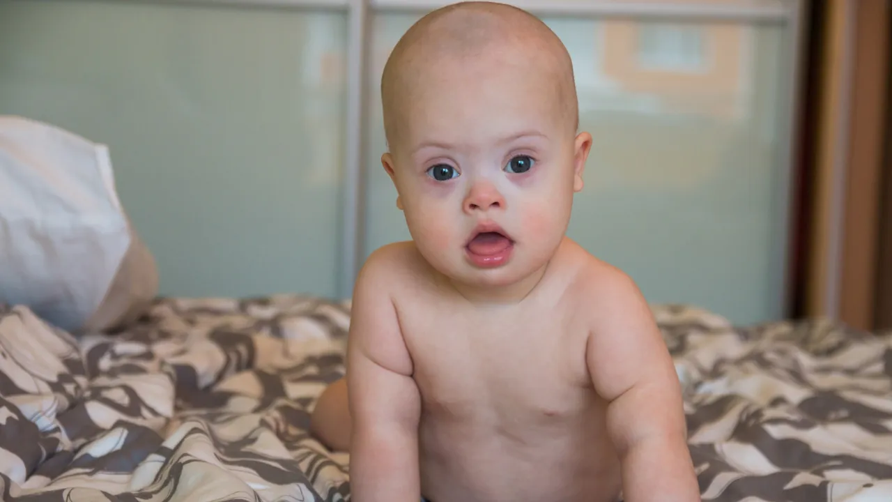 A baby sits on a bed and looks directly at the camera