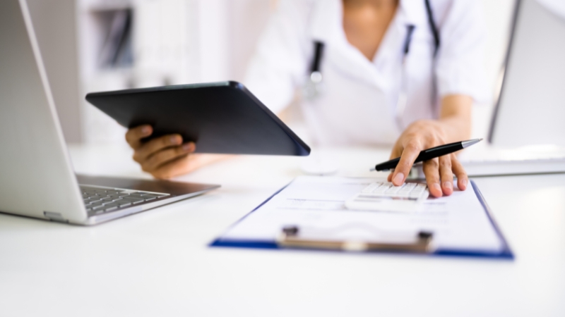 A doctor checks a tablet and inputs data next to a clipboard and laptop to ensure compliance with healthcare regulations