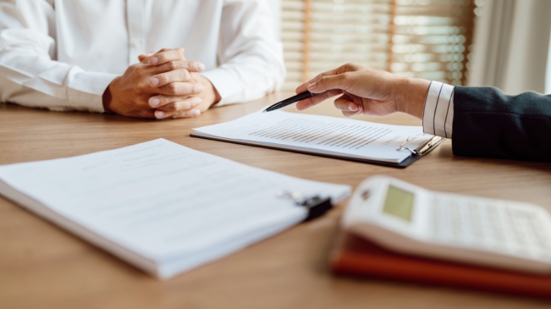 Two people sit across a desk reviewing documents, with a calculator and contract visible during a discussion