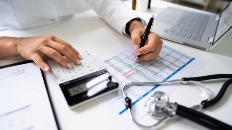 A healthcare professional calculates billing data with a pen and calculator beside a laptop and stethoscope in a private medical practice