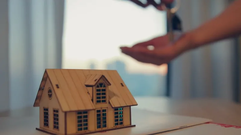 A small wooden house model sits on a table as someone hands over keys, symbolizing rising homeowners insurance rates in the U.S.