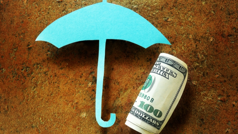 A blue paper umbrella and a rolled $100 bill on a table represent the cost and coverage of umbrella insurance